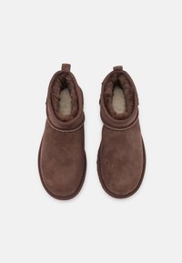 Botas de tobillo de ante marrón con un suave forro de piel sintética, punta redondeada y costuras decorativas alrededor del tobillo. Diseño texturizado y acogedor.