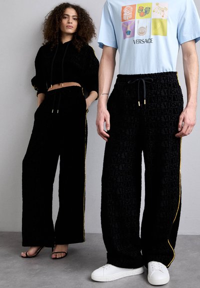 Ensemble coordonné noir texturé avec des rayures latérales jaunes ; hoodie court et pantalon ample. Logo Versace en relief sur l'ensemble. Baskets blanches.