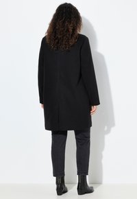 Cappotto di lana nero con taglio dritto, maniche lunghe e una semplice cucitura sul retro. Abbinato a jeans scuri e stivaletti alla caviglia.