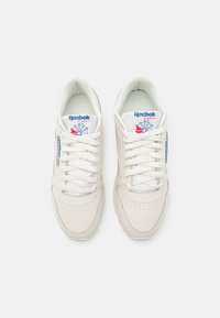 Baltos Reebok sportbačiai su lygiu odiniu paviršiumi, apibrėžta siūlų linija, paminkštinta apykaklė ir logotipas ant liežuvėlio bei šono. Su raišteliais.