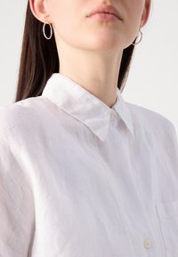 Camisa branca de linho com botão e gola, com um bolso na frente e um corte descontraído. Acessorizada com brincos de argola prateados.