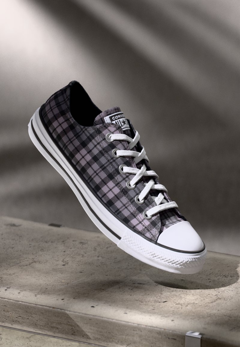 Sneaker Converse a scacchi neri e grigi con lacci e suola bianchi, sospesa sopra una superficie di cemento in una luce morbida e ombreggiata.