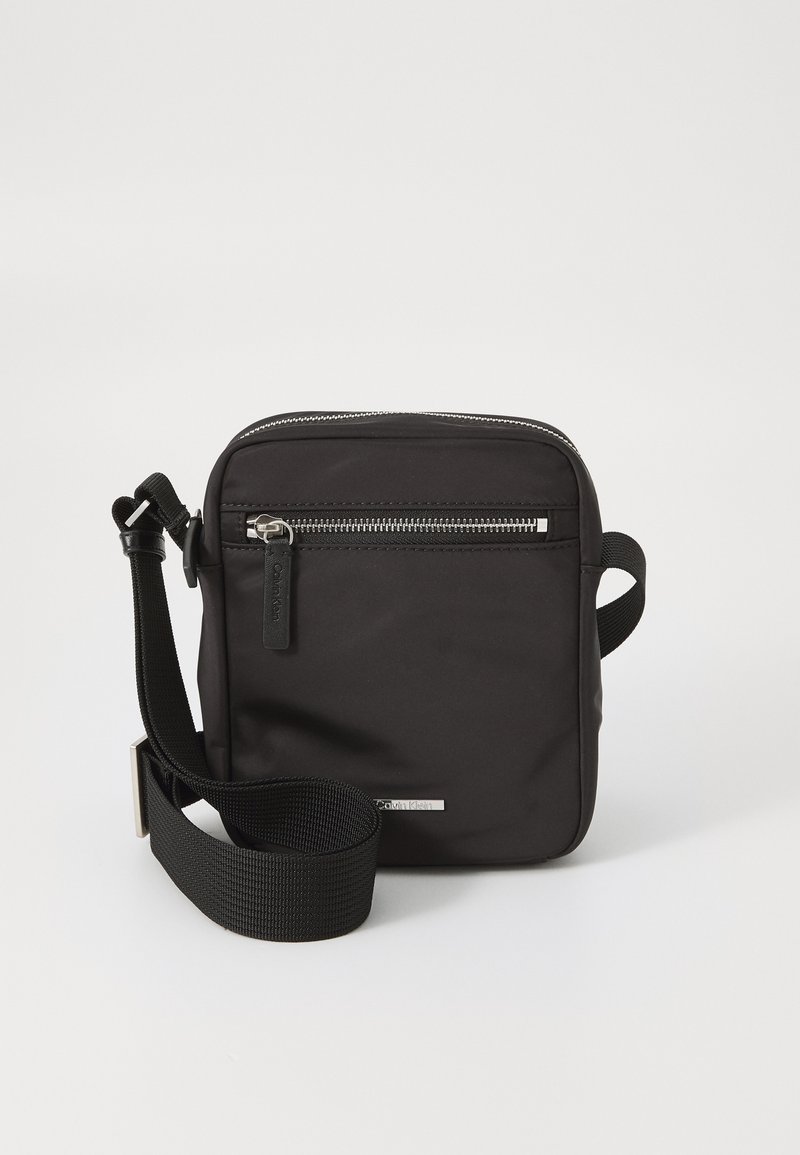 Sac bandoulière noir en nylon résistant. Comprend une poche avant zippée, une sangle ajustable et un accent en métal argenté. Forme carrée.