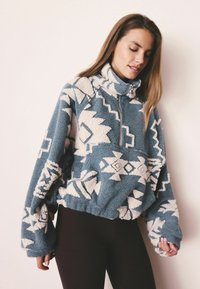 Fleece-pullovari teallin värissä, jossa on valkoisia geometrisiä kuvioita; siinä on puolivetoketju ja elastinen helma; rento istuvuus ja ylimitoitetut hihat.