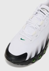 Witte sportieve schoen met een textuur bovenwerk, zwarte neuskap, groene accentkleur en zichtbare luchtkussen in de zool. Vetersluiting.