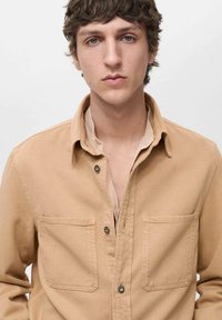 Camicia button-up in cotone beige con due tasche sul petto, colletto a punta e cuciture in vista; indossata sopra una camicia di colore chiaro.