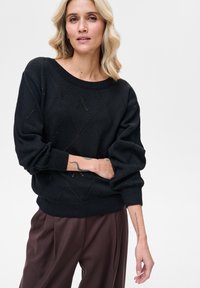 Czarny sweter dziany o luźnym kroju, z rombowymi wycięciami i balonowymi rękawami. W połączeniu z brązowymi spodniami. Gładka tekstura, casualowy design.