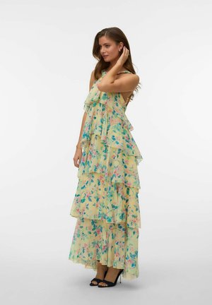 Vrouw staand in een gelaagde, mouwloze gele jurk met blauwe en roze bloemenprint, draagt zwarte hoge hakken met open tenen.