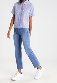 Persona vestida con una camisa de botones de rayas azules y blancas de manga corta, jeans cortos de color azul claro y zapatillas blancas, de pie con las piernas cruzadas.