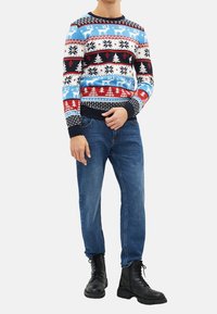 Homme portant un pull festif avec des motifs de rennes et de sapins de Noël, un jean bleu et des bottes noires à lacets sur un fond blanc.
