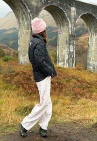 Osoba v růžové čepici, černé bundě a bílých kalhotách kráčí poblíž travnaté plochy s velkým kamenným obloukovým mostem a kopci v pozadí.