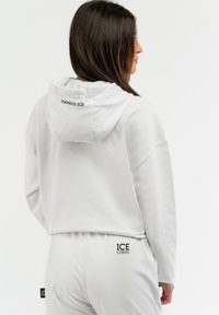 Sudadera blanca cortada con una textura suave y bolsillo tipo canguro, con la impresión "ICE ICEBERG" en la parte de atrás y pantalones de chándal a juego.