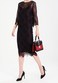Svart spetsklänning med en genomskinlig bodice och trekvartslång ärm, prydd med ett diamantmönster. Kombinerad med en tvåfärgad handväska.