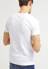 Bílý bavlněný T-shirt se standardním kulatým výstřihem a krátkými rukávy. Zadní pohled ukazuje hladkou látku a minimální detaily. Nosí se s modrými džínami.