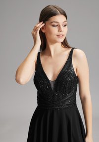 Robe de soirée noire avec un décolleté plongeant, ornée de perles élaborées et de détails en sequins sur le bustier, jupe fluide et lisse.