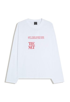 Bela majica z dolgimi rokavi z rdečim napisom »UMETNOST ZA VSE, DA JO DOŽIVIJO IN UŽIVAJO« nad logotipom »THE MET« na sredini sprednjega dela.