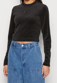 Svart långärmad croppad topp med strukturerat rutmönster och Nike-logotyp på bröstet, kombinerad med högmidjade blå denimbyxor.