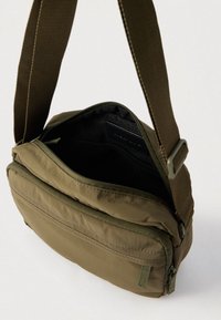 Λαδί τσαντάκι χιαστί με ρυθμιζόμενο λουράκι, μπροστινή τσέπη με φερμουάρ και μερικώς ανοιχτό κύριο διαμέρισμα που δείχνει εσωτερική τσέπη από δίχτυ.