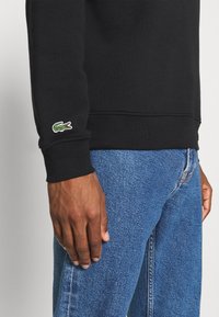 Bras droit et main d'une personne portant un sweat-shirt noir avec un petit logo de crocodile vert sur la manche et un jean bleu en denim.