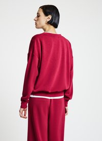 Sweatshirt rouge surdimensionné avec un col ras du cou et des coutures d'épaules tombantes, associé à un pantalon assorti avec une ceinture blanche.