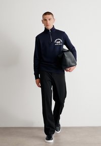 Navy zip-up sweatshirt with white logo detail, paired with black trousers and black sneakers; model carries a black leather bag.
