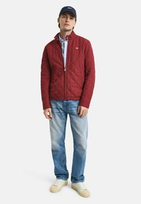 Rode gewatteerde jas met ritssluiting, kraagontwerp, gecombineerd met een lichtblauw shirt, lichtblauwe jeans en witte schoenen. Donkerblauwe pet op het hoofd.