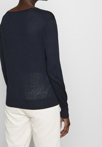 Granatowy sweter dziany z szerokim dekoltem i długimi rękawami, o delikatnej fakturze i ściągaczu u dołu, zestawiony z jasnymi spodniami o lekkiej fakturze.