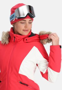 Rode en witte skijas met faux fur afwerking, ritssluiting zakken en zwarte accenten. Gecombineerd met een roze gebreide muts en gespiegelde skibril.