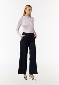 Helllila langärmliges Oberteil mit hohem Kragen und Naht vorne, kombiniert mit weit geschnittenen, dunkelblauen Jeans mit goldenen Knöpfen. Silberne High Heels.