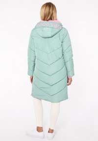 Puffiger mintgrüner langer Mantel mit einer rosa Kapuze, die mit "GOOD VIBES" beschriftet ist. Verfügt über ein Chevron-Quilting und eine lockere Passform. Weiße Leggings und Sneakers.