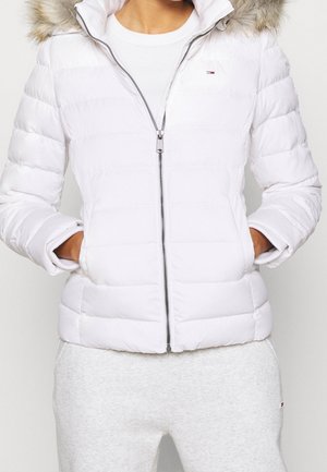 Personne portant une doudoune blanche avec capuche bordée de fourrure, zippée à mi-hauteur, les mains dans les poches, portée avec un pantalon de survêtement gris clair.