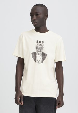 Mand iført creme t-shirt med sort-hvid grafik af en næsehorn i formelt jakkesæt og beskyttelseshjelm, stående mod en ensfarvet baggrund.