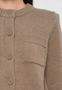 Gros plan d'un cardigan beige en tricot avec des boutons ronds et une petite poche poitrine sur le côté gauche.
