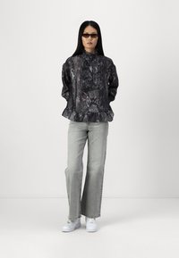 Blusa nera con motivo a serpente e dettagli di volant, abbinata a jeans grigi chiari a gamba larga e sneaker bianche. Il modello indossa occhiali da sole piccoli.