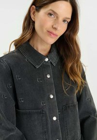 Chaqueta de mezclilla negra con cuello, botones de presión plateados, dos bolsillos frontales y un patrón de logo en relieve en toda la tela.