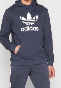 Sudadera color azul marino de mezcla de algodón, con un gran logo blanco de Adidas y un bolsillo frontal. Incluye una capucha con cordón y puños con ribete.