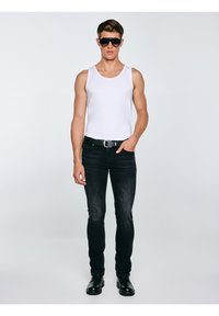 Fehér ujjatlan pólót, fekete farmert, fekete övet, fekete csizmát viselő férfi, nagy fekete napszemüveggel, egyik kezét zsebbe dugva áll.