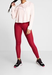 Vrouw in rode legging, zwart-roze Puma sneakers, en een lichtroze lange mouwen top die met één hand haar vlecht vasthoudt.