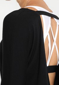 Svart oversized topp med öppen bakdesign, som visar vita korsade band under, tillverkad av mjukt tyg med en slät textur.