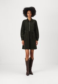Rochie din catifea verde închis, cu fermoar frontal, talie strânsă și două buzunare pe piept. Asociază-o cu cizme cowboy maro pentru contrast.
