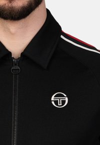 Čierna bunda na zips s golierom, so bielymi a červenými pruhmi na pleciach a bielym logom na ľavej hrudi. Mäkko štruktúrovaná látka.