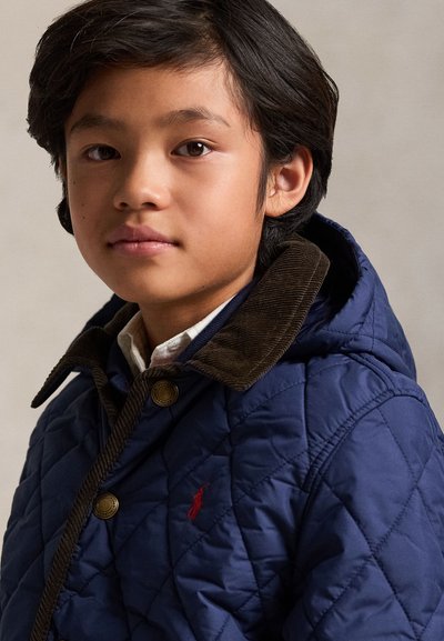 Jeune garçon aux cheveux foncés portant une veste matelassée bleu marine avec un col en velours marron et une chemise blanche en dessous, tourné légèrement vers la gauche.