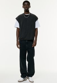 Zwarte oversized mouwloze shirt gelaagd over een wit T-shirt, gecombineerd met losse zwarte spijkerbroek en witte sneakers met zwarte strepen.