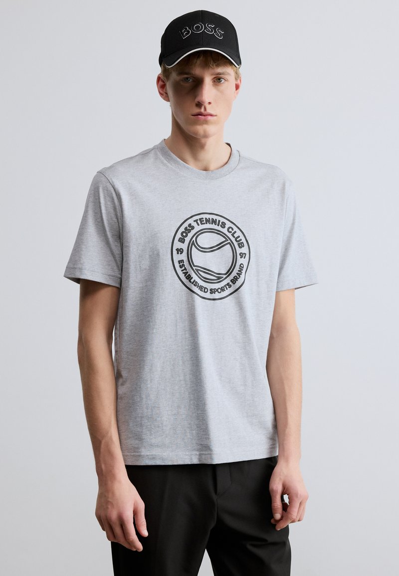 Pilka medvilninė marškinėlė su apskritu atspausdintu logotipu ir tekstu, derinama su juoda „BOSS“ ženkleliu pažymėta kepure ir lygios audinių tekstūros.