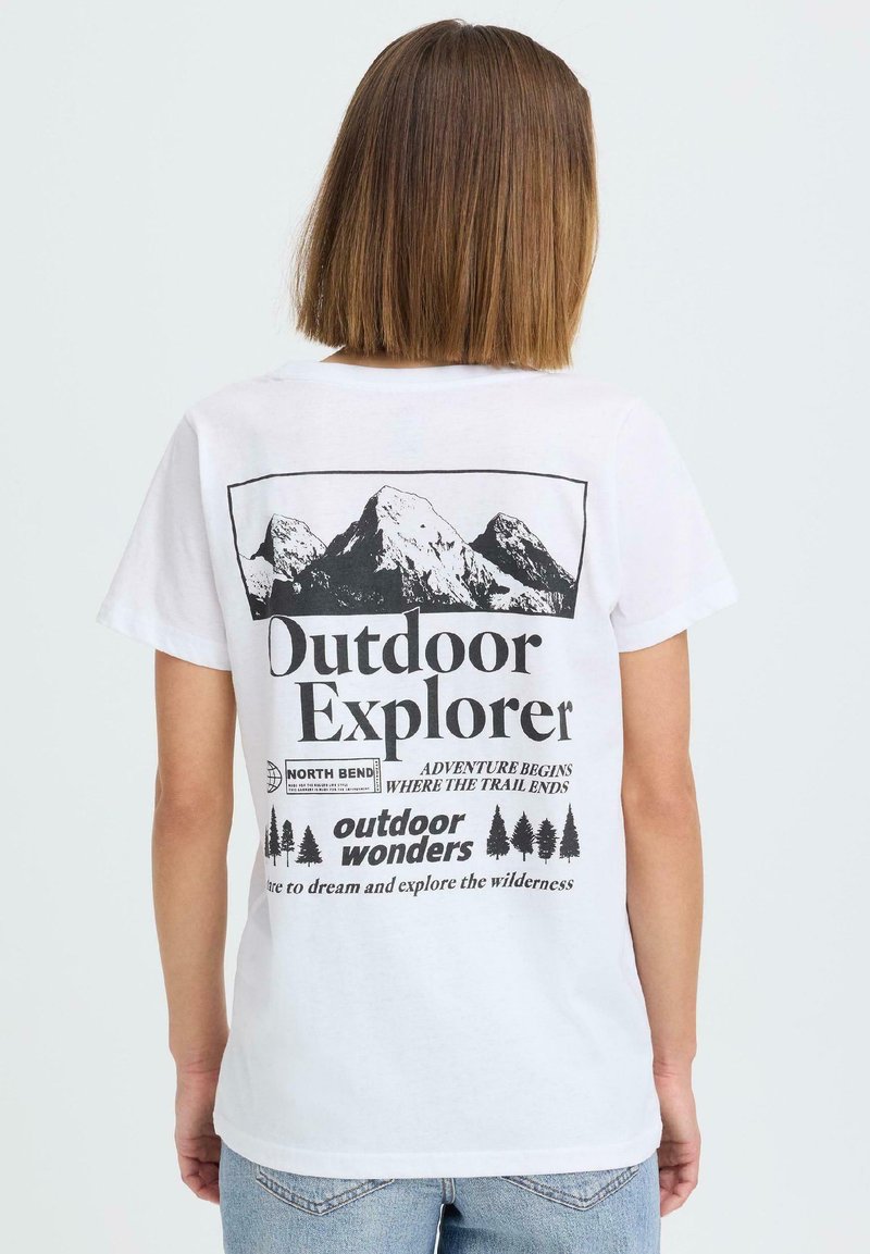 Person med skulderlangt hår iført en hvit T-skjorte med svarte fjell- og treillustrasjoner og utendørstema tekst på baksiden.