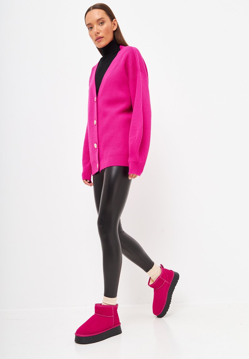 Cardigan ribbed roz intensie, cu nasturi mari, purtat peste un pulover cu guler înalt negru, asortat cu colanți negri strălucitori și bocanci roz până la gleznă.