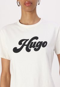T-shirt en coton blanc avec un grand logo cursif noir "Hugo" sur le devant et un col rond classique. Les manches courtes sont de coupe décontractée.