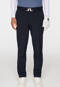 Marineblauwe golfbroek van rekstof, met een slim fit, voorzien van een zwarte riem en zilverkleurige hardware, gecombineerd met witte sneakers en een handschoen.