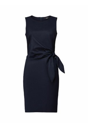 Kobieta w granatowej sukience bez rękawów z wiązaniem po bokach, długie falowane brązowe włosy, srebrne pierścionki i bransoletka.