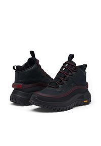 Chaussures de randonnée montantes noires avec des accents rouges, tige texturée, lacets et semelle en caoutchouc épaisse. Présente le logo "SOREL" sur le côté.